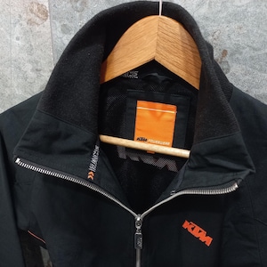 May include: Black zip-up jacket with a high collar and orange accents. The jacket features the KTM logo in orange on the chest and the text "RACE" on the sleeve. The jacket is hanging on a wooden hanger.