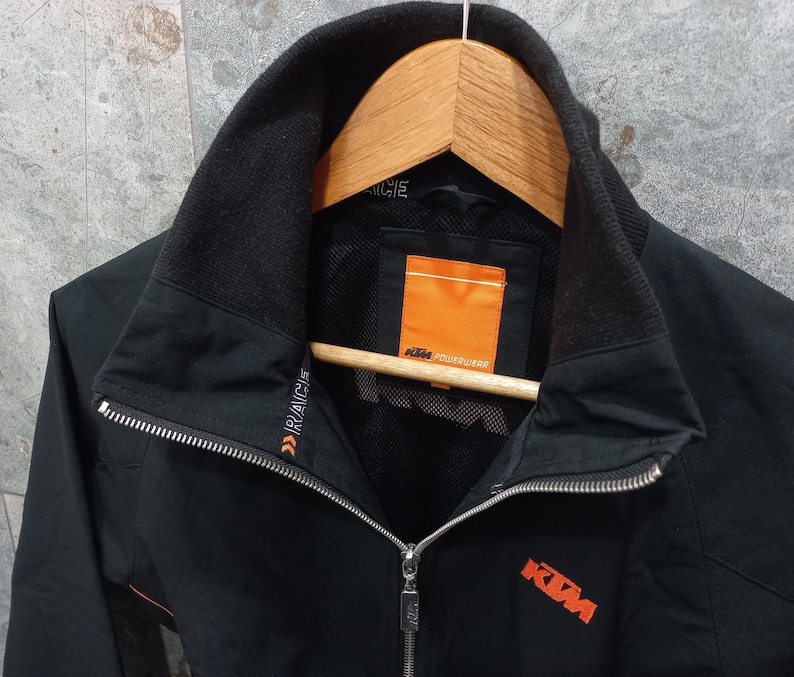 May include: Black zip-up jacket with a high collar and orange accents. The jacket features the KTM logo in orange on the chest and the text "RACE" on the sleeve. The jacket is hanging on a wooden hanger.