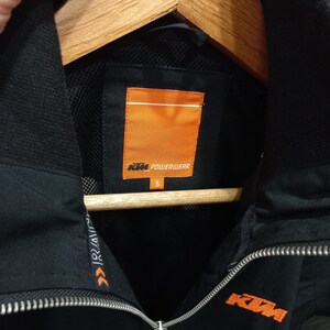 May include: Black KTM Powerwear jacket with a full zipper and a high collar. The jacket features the KTM logo in orange on the chest and the Powerwear logo on the inside. The jacket is size small.