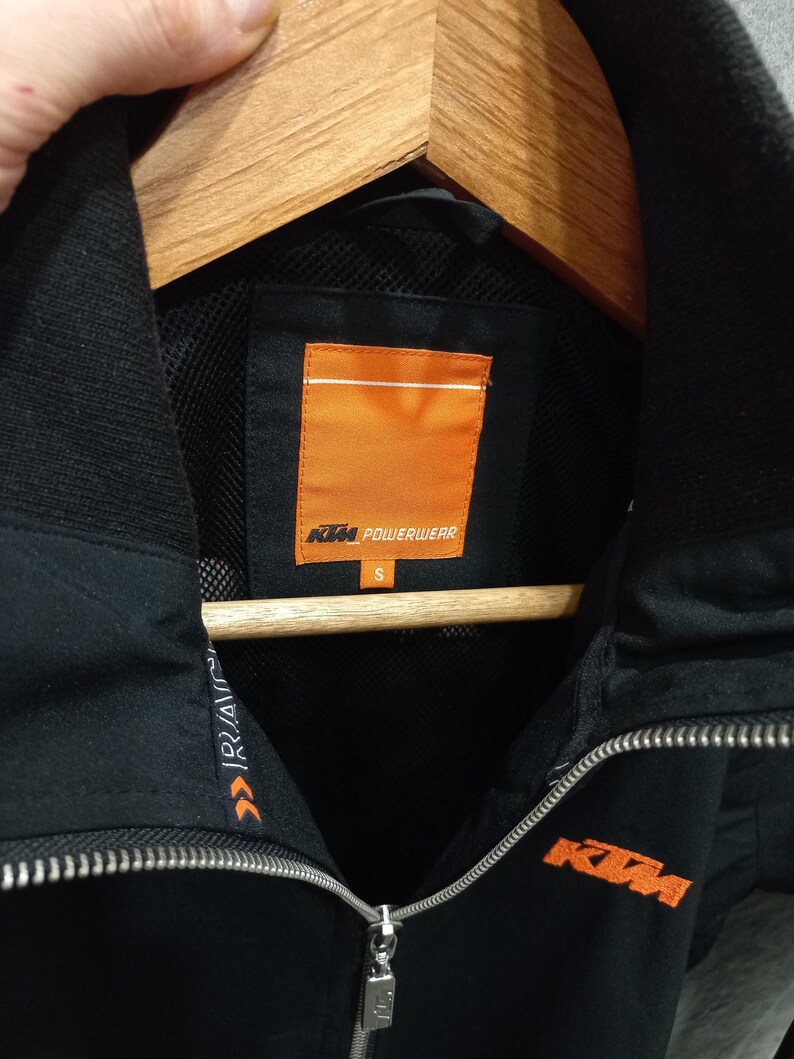 May include: Black KTM Powerwear jacket with a full zipper and a high collar. The jacket features the KTM logo in orange on the chest and the Powerwear logo on the inside. The jacket is size small.
