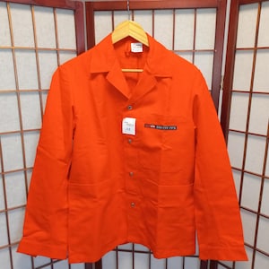 May include: Bright orange work jacket with a button-down front, two front pockets, and a small patch on the right chest. The jacket is hanging on a wooden hanger. The jacket has the text "SBB CFF FFS" on the patch.