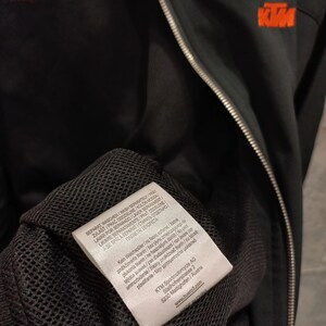 May include: Black KTM motorcycle jacket with a zipper closure and orange logo on the chest. The jacket has a mesh lining and a tag with washing instructions and the KTM Sportmotorcycle AG logo.