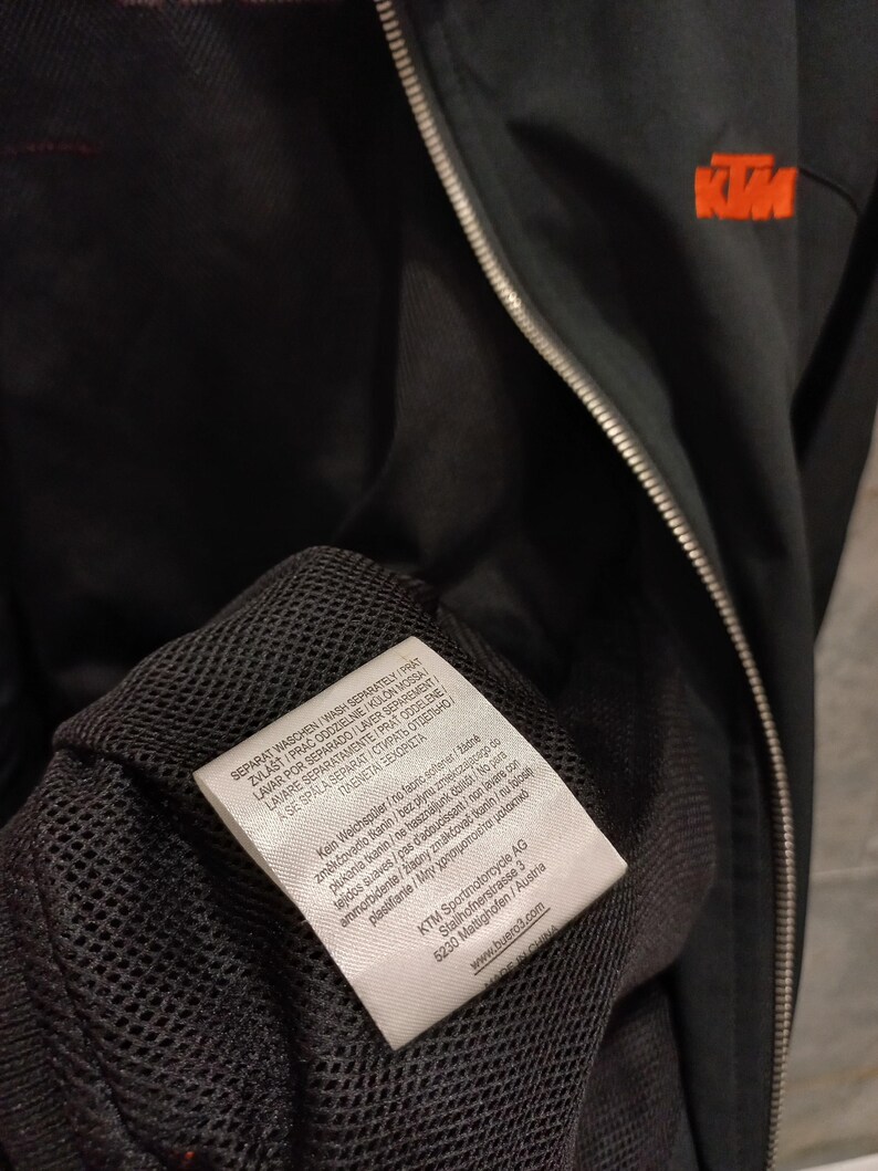 May include: Black KTM motorcycle jacket with a zipper closure and orange logo on the chest. The jacket has a mesh lining and a tag with washing instructions and the KTM Sportmotorcycle AG logo.