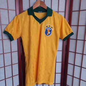 以下が含まれることがあります： 襟と袖にグリーンのトリムが付いた黄色のサッカーユニフォーム。ユニフォームはVネックのデザインで、星と「CBF」の文字が入った紋章が付いています。シャツは木製のハンガーに吊るされています。