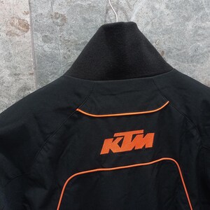 May include: Black jacket with orange trim and the KTM logo on the back. The jacket has a black collar and a zipper closure. The KTM logo is in orange. The jacket is hanging on a hanger.