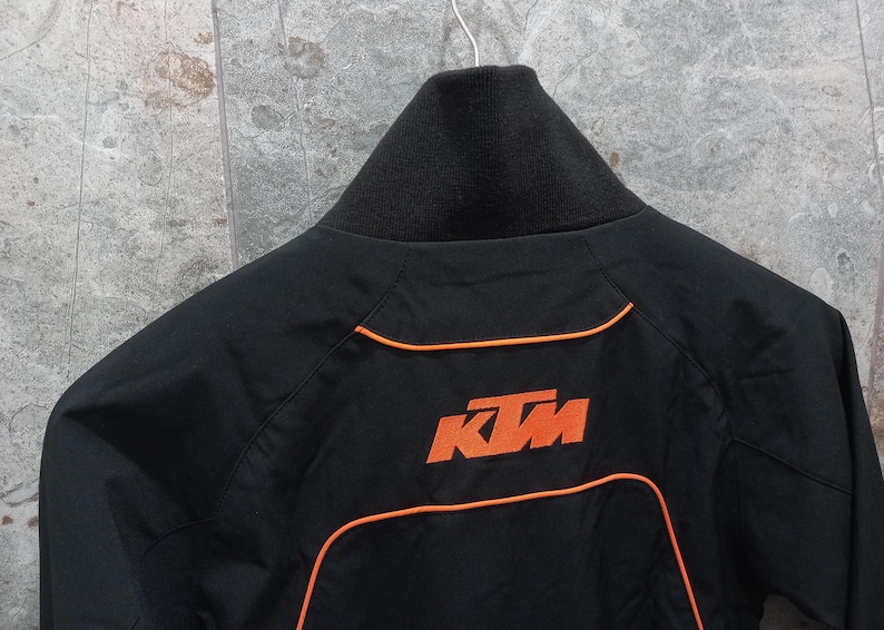 May include: Black jacket with orange trim and the KTM logo on the back. The jacket has a black collar and a zipper closure. The KTM logo is in orange. The jacket is hanging on a hanger.