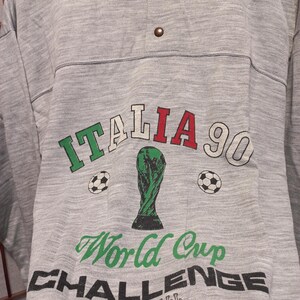 Puede incluir: Una camiseta gris de manga larga con cuello rojo y tapeta abotonada. La camiseta presenta el texto "ITALIA 90" en verde, rojo y blanco, junto con un trofeo verde y balones de f&uacute;tbol. Tambi&eacute;n est&aacute;n impresas las palabras "World Cup Challenge Football".