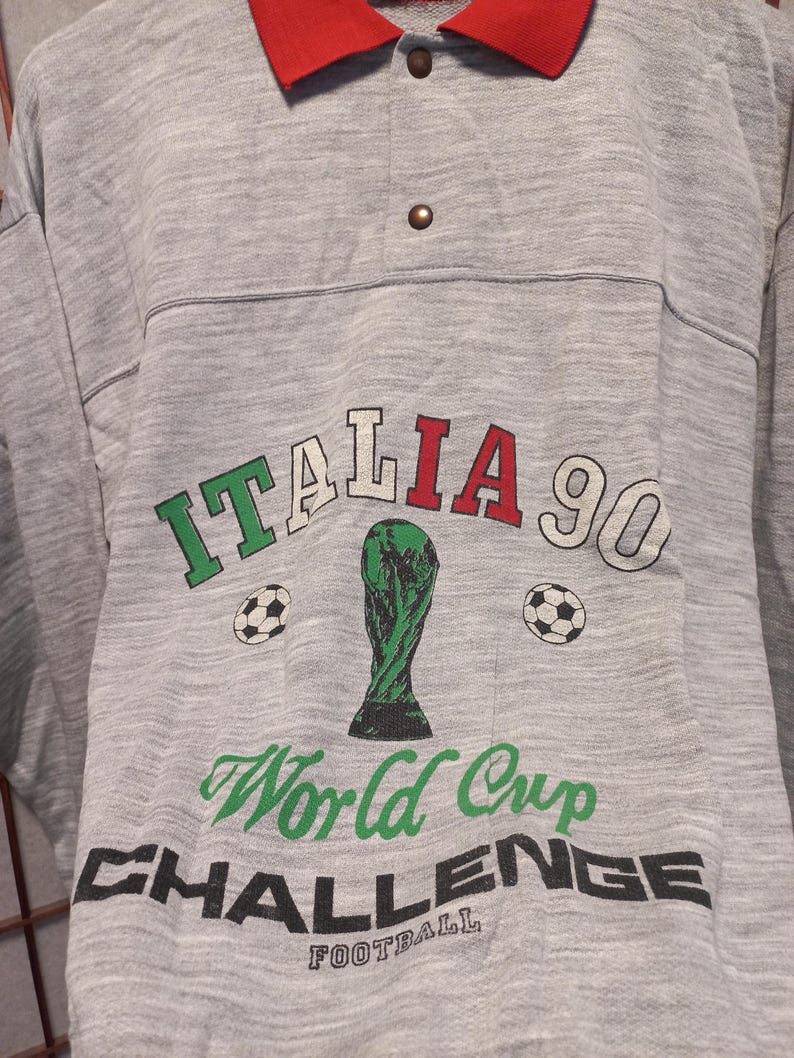 Puede incluir: Una camiseta gris de manga larga con cuello rojo y tapeta abotonada. La camiseta presenta el texto "ITALIA 90" en verde, rojo y blanco, junto con un trofeo verde y balones de f&uacute;tbol. Tambi&eacute;n est&aacute;n impresas las palabras "World Cup Challenge Football".