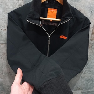 May include: Black zip-up jacket with a stand-up collar and ribbed cuffs. The jacket features the KTM logo in orange on the chest. The jacket is hanging on a wooden hanger.