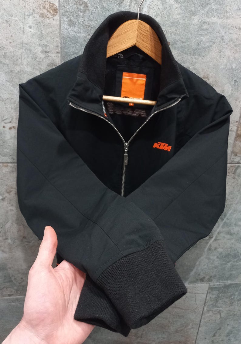 May include: Black zip-up jacket with a stand-up collar and ribbed cuffs. The jacket features the KTM logo in orange on the chest. The jacket is hanging on a wooden hanger.