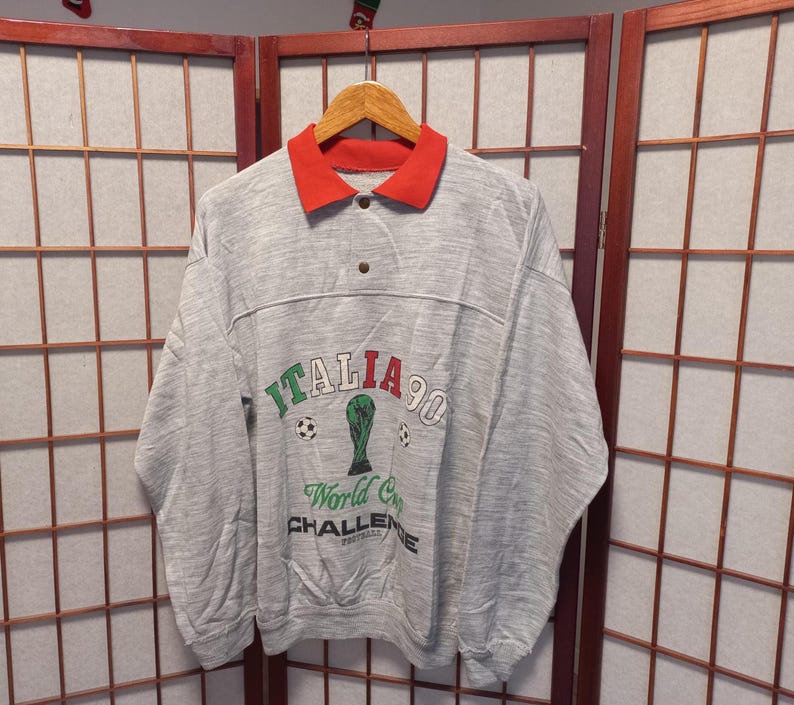 Puede incluir: Una sudadera gris con cuello rojo y el texto "Italia 90 World Cup Challenge Football". El gr&aacute;fico incluye un bal&oacute;n de f&uacute;tbol y un trofeo. La sudadera est&aacute; colgada en una percha de madera.