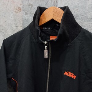 May include: Black zip-up jacket with a high collar and orange accents. The jacket features the KTM logo in orange on the front. The jacket is hanging on a wooden hanger.