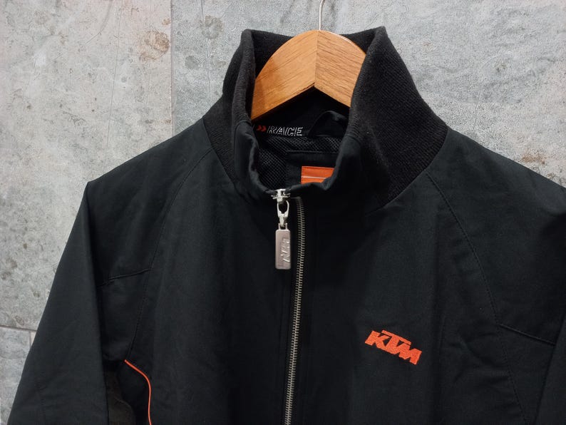 May include: Black zip-up jacket with a high collar and orange accents. The jacket features the KTM logo in orange on the front. The jacket is hanging on a wooden hanger.
