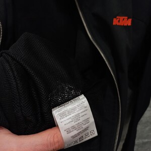 May include: Black zip-up jacket with the KTM logo in orange on the chest. The jacket has a silver zipper and a tag that says "Designed by b&uuml;ro3". The jacket is made of polyester and elastane.