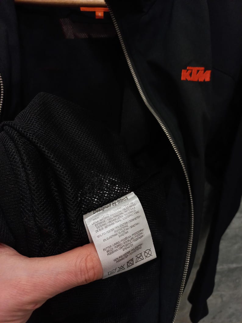May include: Black zip-up jacket with the KTM logo in orange on the chest. The jacket has a silver zipper and a tag that says "Designed by b&uuml;ro3". The jacket is made of polyester and elastane.
