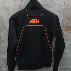May include: Black zip-up jacket with orange trim and the KTM logo on the back. The jacket has a high collar and a sporty design. The KTM logo is in orange. The jacket is hanging on a metal hanger.