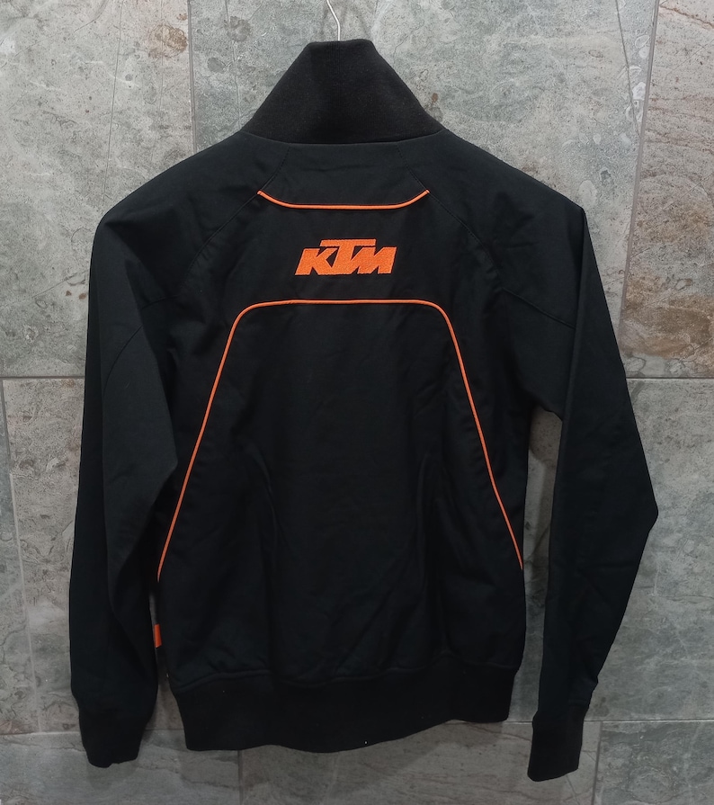May include: Black zip-up jacket with orange trim and the KTM logo on the back. The jacket has a high collar and a sporty design. The KTM logo is in orange. The jacket is hanging on a metal hanger.