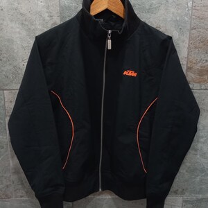 May include: Black zip-up jacket with orange accent lines and the KTM logo in orange on the chest. The jacket has a high collar and a full-length zipper. The jacket is hanging on a wooden hanger.