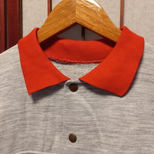 Puede incluir: Primer plano de una sudadera gris con cuello rojo brillante y dos botones marrones. La sudadera est&aacute; colgada de una percha de madera. El cuello es de un color que contrasta con la tela gris.