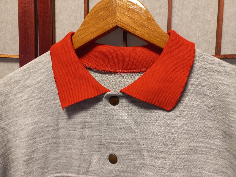 Puede incluir: Primer plano de una sudadera gris con cuello rojo brillante y dos botones marrones. La sudadera est&aacute; colgada de una percha de madera. El cuello es de un color que contrasta con la tela gris.