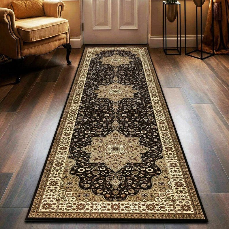 May include: A long, rectangular rug with an ornate, traditional design. The rug features a dark brown background with beige and cream-colored floral patterns and borders. The rug is placed on a hardwood floor.