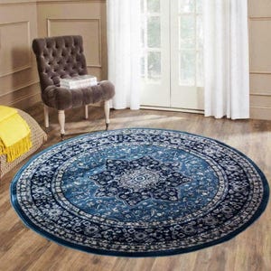 May include: A round, blue area rug with an intricate floral pattern. The rug features a central medallion design and a border with repeating motifs. The rug is placed on a hardwood floor, with a tufted chair and a window in the background.
