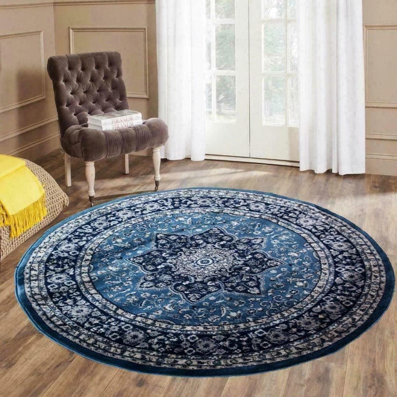 May include: A round, blue area rug with an intricate floral pattern. The rug features a central medallion design and a border with repeating motifs. The rug is placed on a hardwood floor, with a tufted chair and a window in the background.