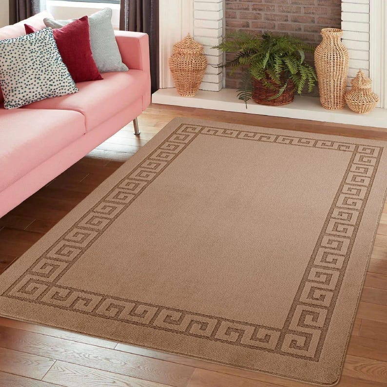 May include: A rectangular beige area rug with a Greek key border design. The rug is placed on a hardwood floor in a living room setting. The border is a darker shade of brown. The rug is a neutral color and would complement various decor styles.