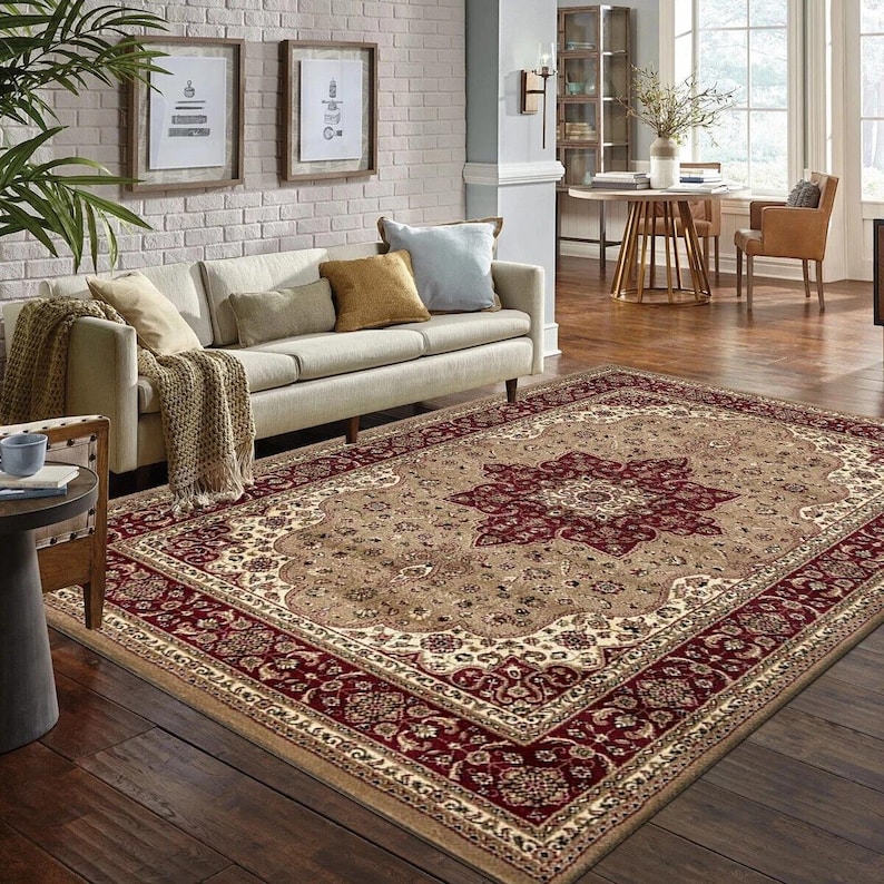 May include: A large, rectangular area rug with an ornate, traditional design. The rug features a central medallion in deep red, surrounded by a beige field and a red border. The rug is placed in a living room setting with a sofa and decorative pillows.