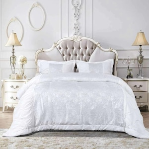 May include: A white floral patterned bed set, comprising a duvet cover, pillows, and sheets. The bed is in a room with white walls, two bedside tables, and lamps. The headboard is upholstered in a light grey fabric. The room has a classic design.