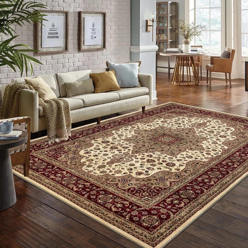 May include: A large rectangular area rug with an ornate, traditional design. The rug features a cream-colored central field with a detailed floral pattern in shades of brown and beige. A deep red border frames the design, adding contrast.