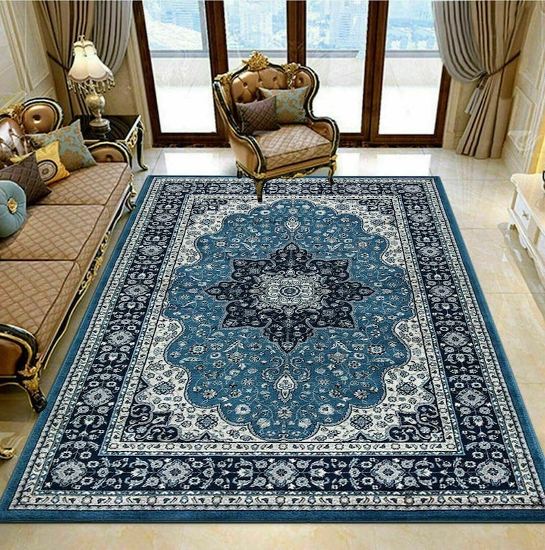 May include: A rectangular area rug with a central medallion design in shades of blue and white. The rug features an ornate border with floral and geometric patterns. The rug is placed on a tiled floor, and is surrounded by furniture.