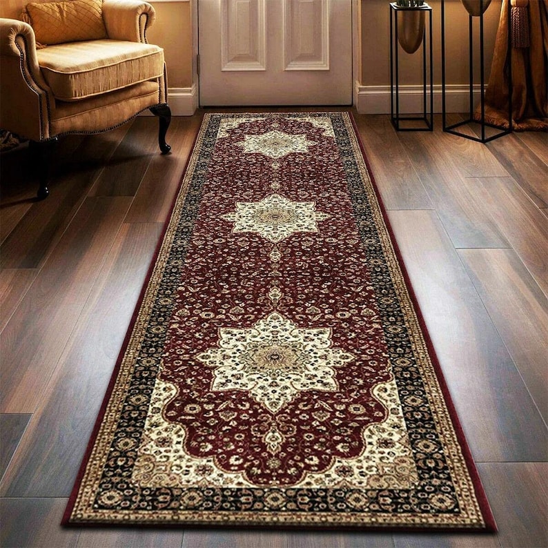 May include: A long, rectangular runner rug with a deep red background and intricate floral patterns in cream, black, and gold. The rug features a central medallion design and borders with detailed motifs, suitable for a hallway or entryway.