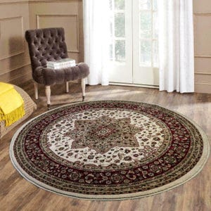 May include: A round area rug with a central floral medallion design in cream, burgundy, and olive green. The rug has a detailed border and is placed on a hardwood floor. A tufted chair and a window are in the background.
