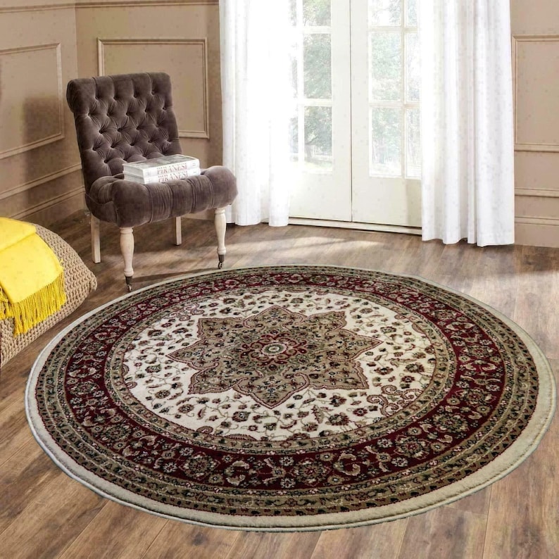 May include: A round area rug with a central floral medallion design in cream, burgundy, and olive green. The rug has a detailed border and is placed on a hardwood floor. A tufted chair and a window are in the background.