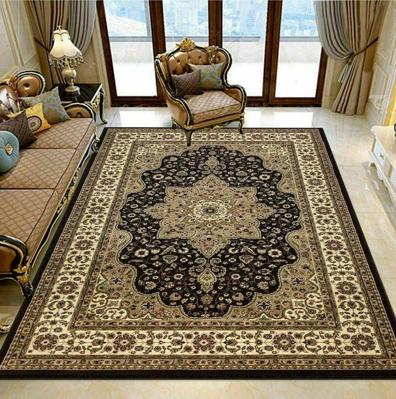 May include: A large rectangular area rug with an ornate floral design in black, beige, and brown. The rug features a central medallion and a detailed border, adding a touch of elegance to the room. The rug is placed on a tiled floor.