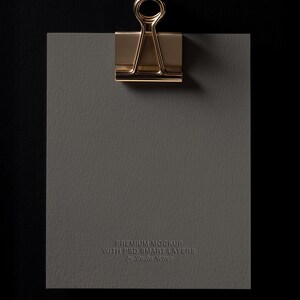 May include: A gold-colored binder clip holds a gray textured paper against a black background. The paper has the text "PREMIUM MOCKUP WITH PSD SMART LAYERS by Studio Netter" embossed on it.