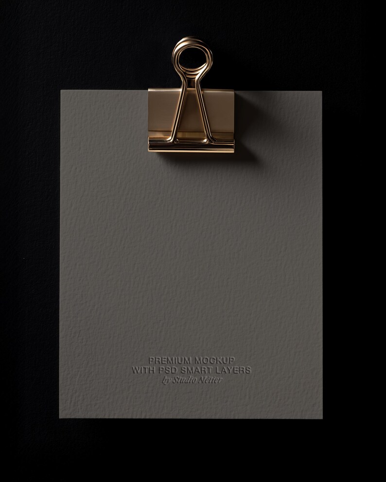 May include: A gold-colored binder clip holds a gray textured paper against a black background. The paper has the text "PREMIUM MOCKUP WITH PSD SMART LAYERS by Studio Netter" embossed on it.