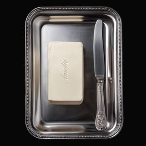Könnte beinhalten: Ein silbernes rechteckiges Serviertablett mit einem Perlenrand enthält einen rechteckigen Butterblock mit dem eingeprägten Namen "Amélie". Ein silbernes Buttermesser liegt auf dem Tablett.