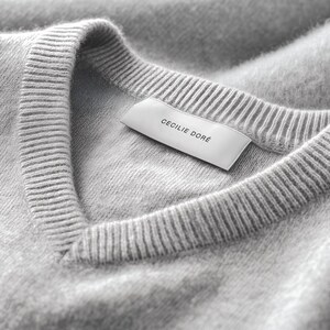 May include: Close-up of a light gray V-neck sweater. The sweater has a ribbed neckline and a label that reads "CECILIE DORÉ". The fabric appears soft and slightly textured, with subtle shading.