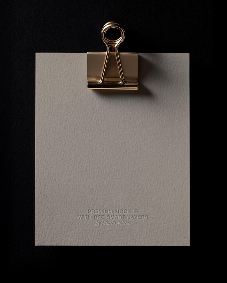 May include: A gold-colored binder clip holding a textured, light gray paper against a black background. The paper has the embossed text "PREMIUM MOCKUP WITH PSD SMART LAYERS by Studio Netter."