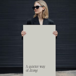 Könnte beinhalten: Eine Person mit Sonnenbrille und schwarzem Blazer hält ein rechteckiges Schild mit dem Text "A quieter way of living." Das Schild ist hellbeige. Der Hintergrund ist eine dunkelgraue, strukturierte Wand.