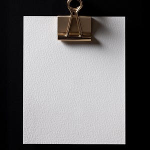 May include: A white textured paper sheet held by a gold-colored binder clip against a black background. The clip is centered at the top, casting a shadow. The paper has a slightly rough surface.
