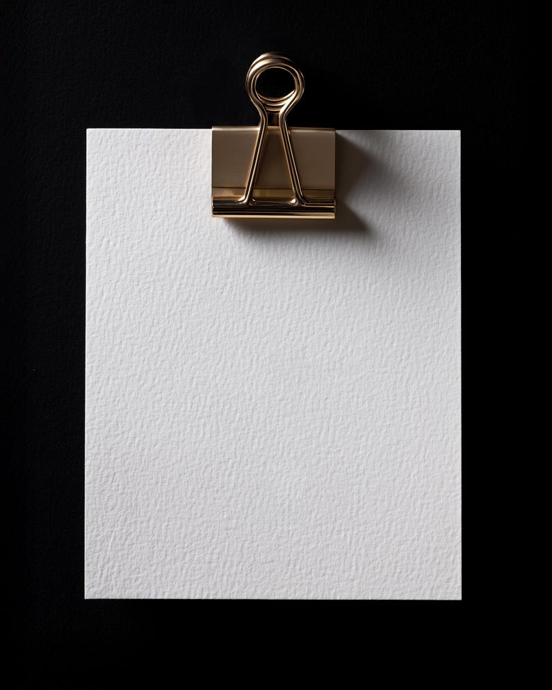 May include: A white textured paper sheet held by a gold-colored binder clip against a black background. The clip is centered at the top, casting a shadow. The paper has a slightly rough surface.