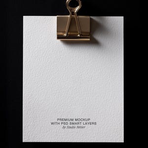 May include: A white textured paper sheet held by a gold-colored binder clip against a black background. The paper has the text "PREMIUM MOCKUP WITH PSD SMART LAYERS by Studio M&eacute;tier" printed at the bottom.