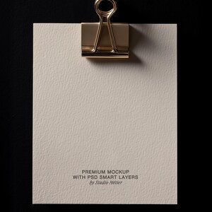 May include: A gold-colored binder clip holding a textured, off-white paper against a black background. The paper has the text "PREMIUM MOCKUP WITH PSD SMART LAYERS by Studio M&eacute;tier" printed at the bottom.