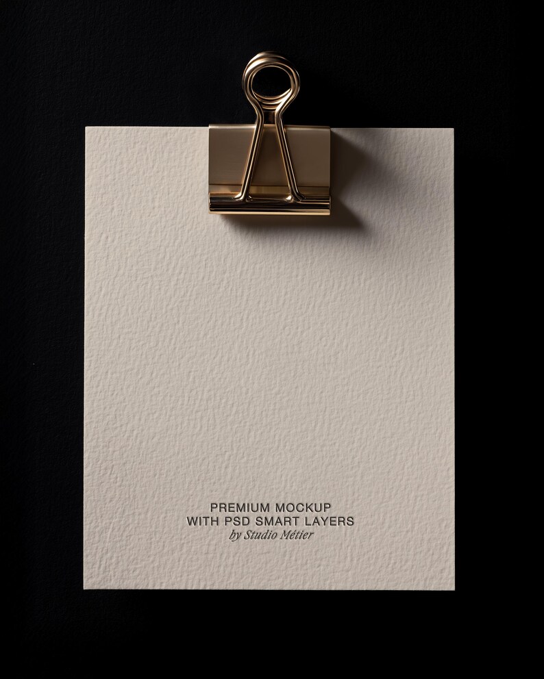 May include: A gold-colored binder clip holding a textured, off-white paper against a black background. The paper has the text "PREMIUM MOCKUP WITH PSD SMART LAYERS by Studio M&eacute;tier" printed at the bottom.
