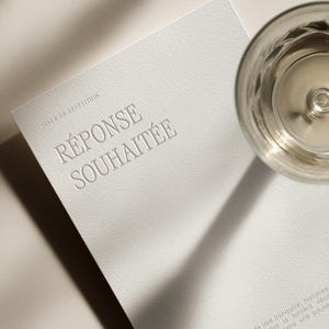 May include: A white card with embossed text reading "RÉPONSE SOUHAITÉE" and "DINER DE RÉPÉTITION". A glass of liquid sits to the right. The card has a textured surface and is angled towards the viewer.