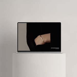 Op de afbeelding: Een zilveren laptop op een witte sokkel. Het scherm toont een zwarte achtergrond met een gouden armband om een pols. De tekst "PSD MACBOOK MOCKUP" is zichtbaar op het scherm.