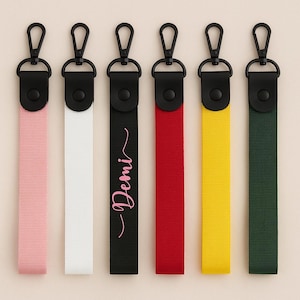 Op de afbeelding: Zes sleutelhangers met zwarte hardware en gekleurde bandjes. De bandjes zijn roze, wit, zwart met de naam "Demi", rood, geel en groen. Elke sleutelhanger heeft een zwarte karabijnhaak voor eenvoudige bevestiging.